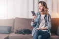 Young woman in denim shirt sitting at home on couch and using smartphone,nearby lies cat. Girl uses digital gadget.