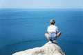 A young woman in denim shorts and a white T-shirt sits on a cliff against the backdrop of the sea and blue sky on a sunny day. Royalty Free Stock Photo