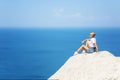 A young woman in denim shorts and a white T-shirt sits on a cliff against the backdrop of the sea and blue sky on a sunny day. Royalty Free Stock Photo