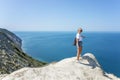 A young woman in denim shorts and a white T-shirt stands on a cliff against the backdrop of the sea and blue sky on a sunny day. Royalty Free Stock Photo