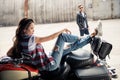 Young woman in denim vest sitting on motorcycle and stylish man in sunglasses standing behind Royalty Free Stock Photo