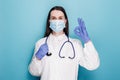 Young woman doctor in medical mask and white coat, rubber gloves, ensure everything alright, show okay gesture