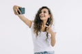 Young woman doing selfie shot pointing index finger on mobile phone showing making stick tongue out sign isolated on white Royalty Free Stock Photo