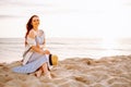 Young woman in a dress holding straw hat and sitting alone on empty sand beach at sunset sea shoreand smiling. Girl looking to the Royalty Free Stock Photo