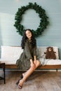 A young woman in a dress is sitting on a white sofa against the background of a large Christmas wreath on the blue wooden wall in Royalty Free Stock Photo