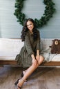 Young woman in a dress is sitting on a white sofa against the background of a large Christmas wreath on the blue wooden wall in Royalty Free Stock Photo