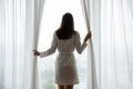 Young woman dressed in bathrobe opening curtains on picture window