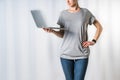 Young woman, dressed in gray t-shirt and blue jeans, standing on light gray background and holding laptop. Royalty Free Stock Photo