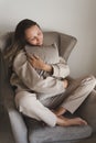 Young woman dressed in grey comfy suit sitting in a cozy armchair and holding a small decorative pillow Royalty Free Stock Photo