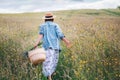 Young Woman dressed jeans jacket and light summer dress walking by the high green grass meadow with basket and wildflowers bouquet Royalty Free Stock Photo