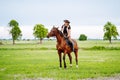 Young woman dressed in riding clothes and hat riding brown horse in green field Royalty Free Stock Photo