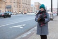 Young woman dressed warm coat, scarf and hat standing roadside, waiting taxi or public transport