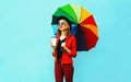 Young woman drinking coffee and holding colorful umbrella walking in red jacket, black hat on blue wall Royalty Free Stock Photo