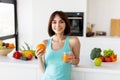 Young woman drinking juice and holding orange, standing near kitchen table full of fruits and smiling at camera Royalty Free Stock Photo