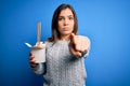 Young woman eating asian noodles from take away box using chopstick over blue background pointing with finger to the camera and to