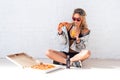 Young woman eating a piece of pizza drinking juice sitting on floor wearing sunglasses Royalty Free Stock Photo