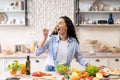 Young Woman Eating Salad At Table With Organic Vegetables, Enjoying Healthy Diet, Standing In Light Kitchen Interior