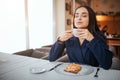 Young woman enjoying her cup of coffee. She sit at table and smell it. Model has cake on plate on table. Royalty Free Stock Photo