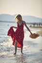 Young woman enjoying her walk on the beach. Beautiful blonde woman in red wavy dress holding straw hat, walking on the shore. Royalty Free Stock Photo