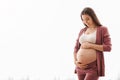 Young woman expecting baby embracing belly while standing near window at home