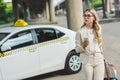 young woman in eyeglasses holding smartphone and looking away while standing near taxi cab