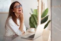 Young woman in eyeglasses laughing while online chatting on computer during the coffee break in cafe. Royalty Free Stock Photo