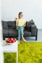 young woman in eyeglasses reading book and holding apple on sofa near table with coffee apples and laptop