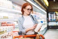 Young woman in eyeglasses and striped shirt with shopping cart thoughtfully looking on bottle of milk in supermarket Royalty Free Stock Photo
