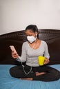 A young woman with face mask sits on the bed and looks at her phone Royalty Free Stock Photo