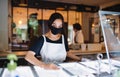 Young woman with face mask working indoors in cafe, disinfecting surfaces.