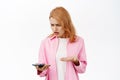 Young woman facing problem on mobile phone, looking frustrated and upset at smartphone, standing over white background Royalty Free Stock Photo