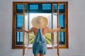 Young woman farmer, dressed in blue jumpsuit shorts, looks out window with open shutters on grape field