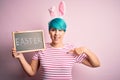 Young woman with fashion blue hair wearing easter rabbit ears and holding blackboard with holiday word with surprise face pointing