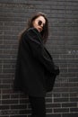 Young woman fashion model in fashionable black blazer in trendy round sunglasses poses near vintage black brick building in street Royalty Free Stock Photo