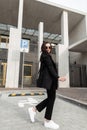 Young woman fashion model in vintage black clothing with handbag in fashion sunglasses walks near modern building in city. Cool Royalty Free Stock Photo