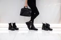Young woman with a fashionable black leather bag in stylish boots in jeans stands near a white wall in a store next to shoes. Royalty Free Stock Photo