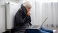 Young woman feeling cold wearing winter hat and coat sitting by the heater nd working on laptop Royalty Free Stock Photo