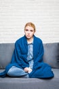 Young woman feeling sick or sad wrapped in cozy blue blanket and sitting on sofa at home Royalty Free Stock Photo