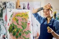 Young woman, female artist in apron looking pleased with her work while creating abstract painting, applying paint on