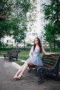 a young woman in a festive short blue corset dress sits gracefully on a wooden bench in a city park in summer