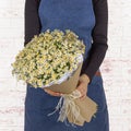 Young woman florist holding big beautiful blossoming bouquet of small white flowers in craft paper cone over white brick Royalty Free Stock Photo