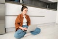 Young woman freelancer, working from home, student sitting with laptop on kitchen floor, wearing headphones, typing on Royalty Free Stock Photo