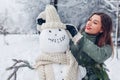 Young woman gives snowman outdoors in snowy winter park her hat, scarf and attaching fir branches in hands. Royalty Free Stock Photo