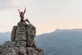 A young woman, glad to have climbed the mountain sits on peak with raised hands and enjoys an amazing view