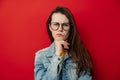 Young woman in glasses curves lips and frowns eyebrows, keeps hand under chin, looks with discontent expression