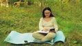 A young woman in glasses on a forest glade working on a tablet and penciling in a notebook