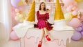 Young woman in glasses, red dress with donut in hand, sitting on table in bright decorative confectionery.