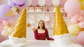 Young woman in glasses, red dress with donut in hand, standing at table in bright decorative confectionery.