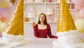 Young woman in glasses, red dress with donut in hand, standing at table in bright decorative confectionery.