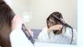 A young woman in glasses straightens her hair with a curling iron in front of a mirror. A girl in a white coat makes a Royalty Free Stock Photo